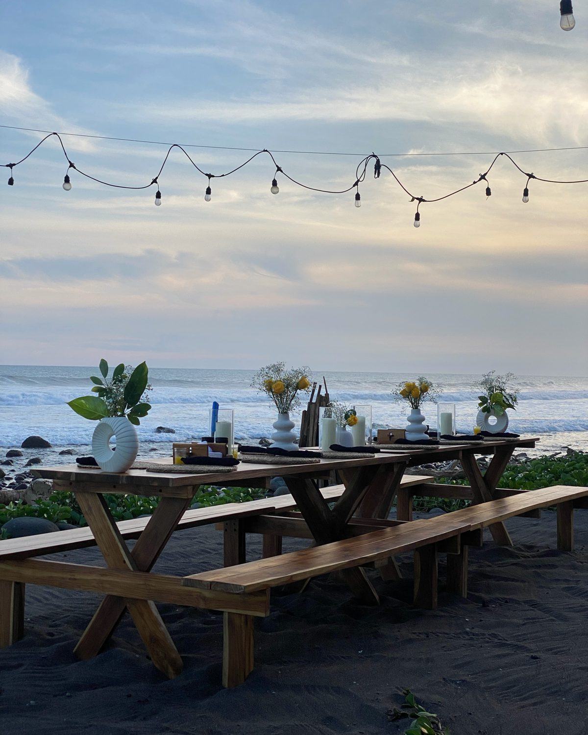 A long table set with candles and flowers on a black-sand beach under string lights