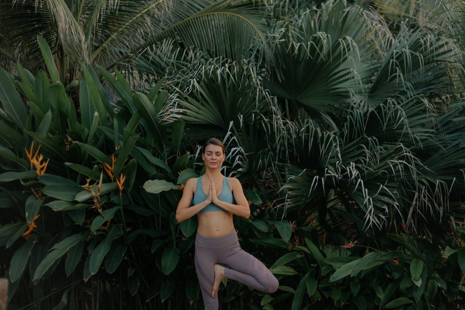 A woman in tree pose with eyes closed, surrounded by tropical palms and heliconia