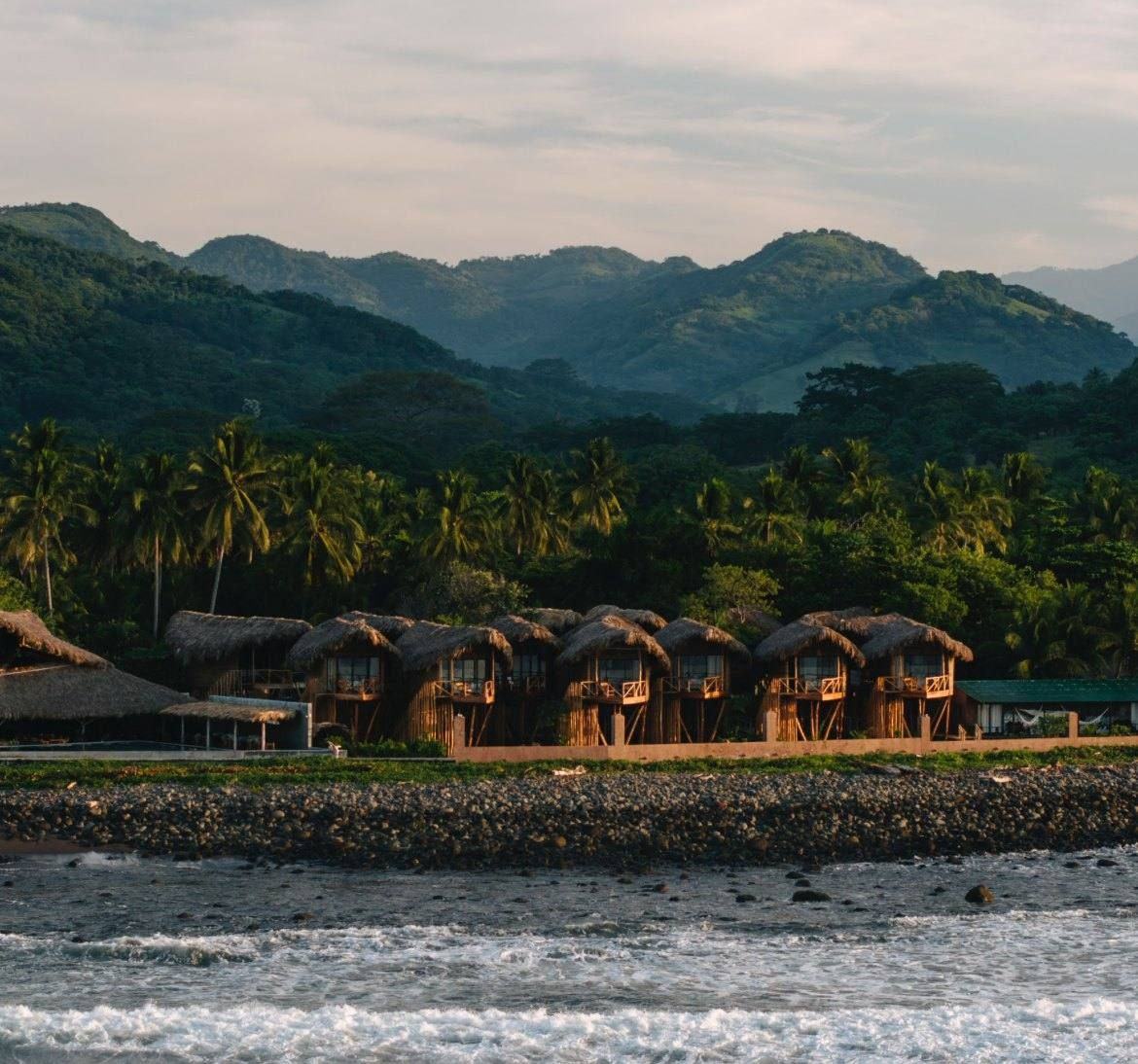 Thatched treehouses on a black-sand beach beneath green mountains at Mizata, El Salvador