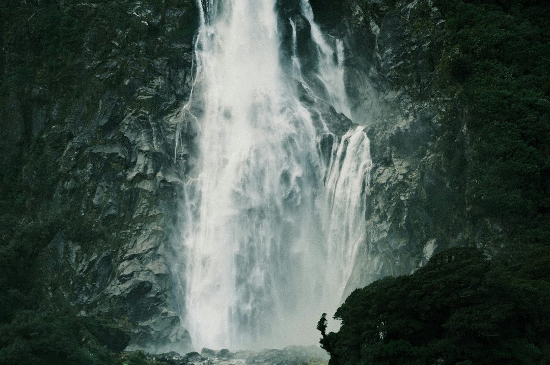 A lone hiker dwarfed by a towering waterfall in a green mountain valley